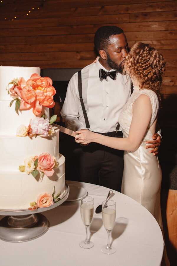 A bride and groom share a kiss while cutting their wedding cake, which is decorated with colorful flowers. Two glasses of champagne sit on the table beside the cake. Fearrington Village