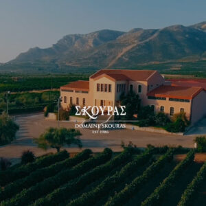 A large, yellow-tan winery building with red roofs stands among vineyards, olive trees, and mountains in the background. White text reads “DOMAIN SKOURAS EST 1986” in both Greek and English at the center. Fearrington Village