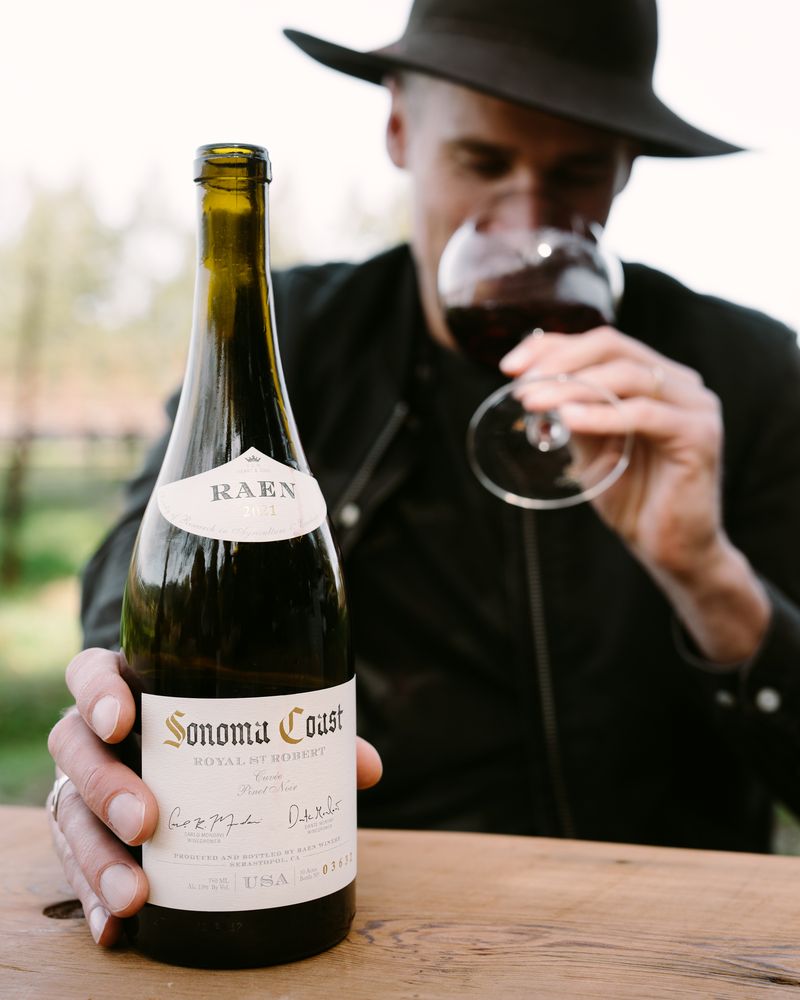 A person in a dark hat and jacket holds a bottle of Sonoma Coast wine labeled "RAEN" on a wooden table, while smelling a glass of red wine outdoors. The background is softly blurred. Fearrington Village