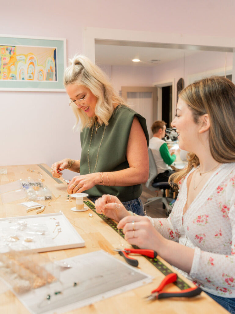 Two women smile and work together making jewelry at a wooden table covered with beads, tools, and supplies in a bright, colorful studio. A person is seated in the background, partially visible. Fearrington Village