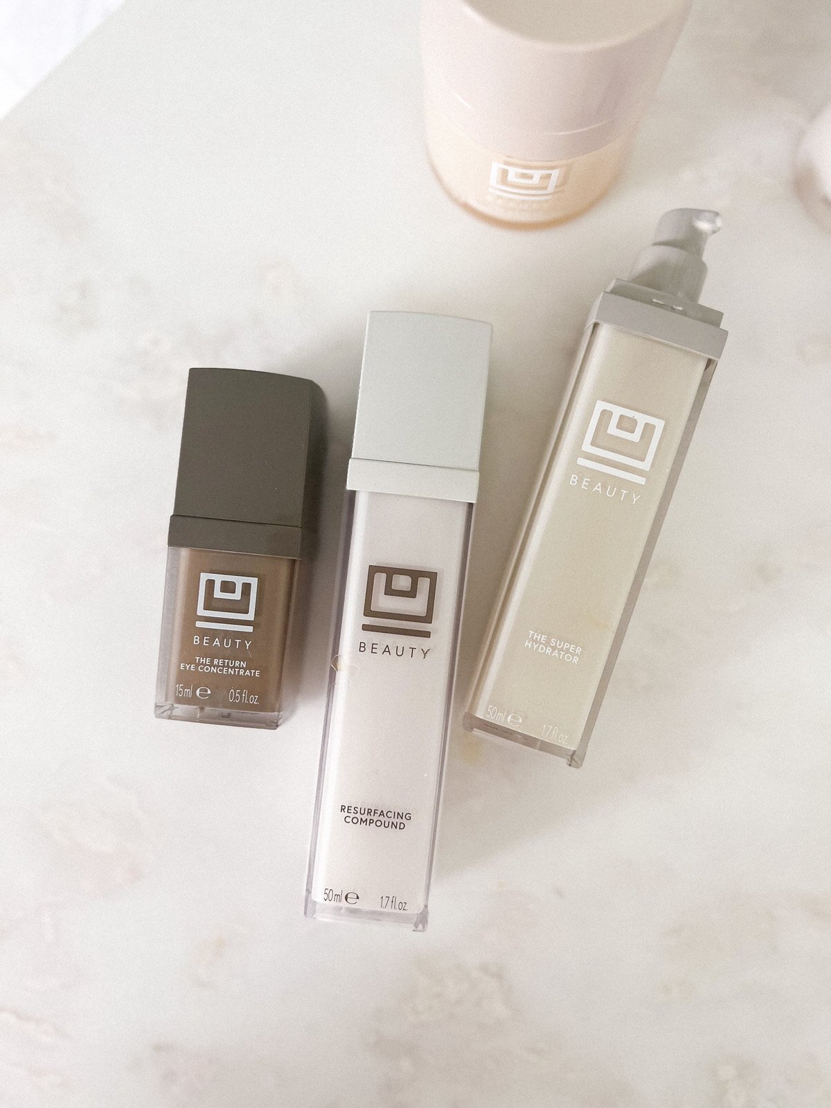 Three skincare products from U Beauty are arranged on a light marble surface. The bottles are sleek and rectangular, featuring the U Beauty logo and labels, including "Resurfacing Compound" and "The Super Hydrator. Fearrington Village