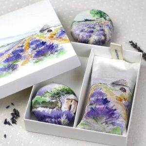 A gift box with a watercolor lavender field design contains a lavender sachet and two round lavender-filled pouches, set on a light polka-dot fabric surface with scattered lavender buds nearby. Fearrington Village