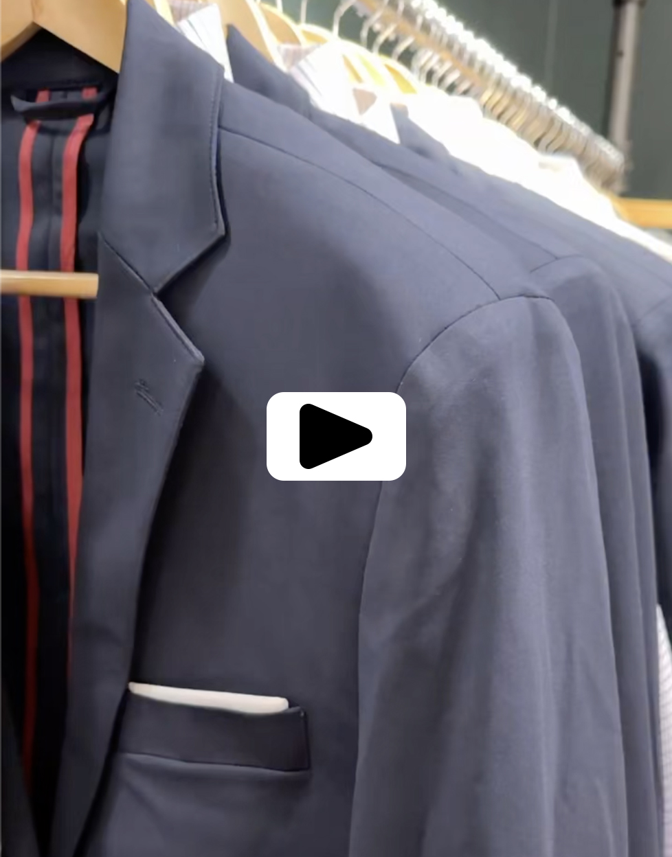 A navy blue suit jacket with a white pocket square hangs on a wooden hanger. Other suit jackets are visible behind it, all hanging on a clothing rack. Fearrington Village