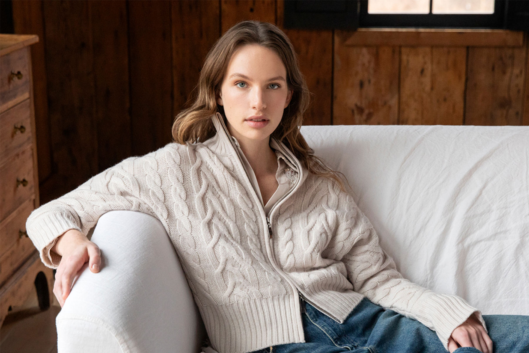 A woman with wavy light brown hair sits on a white couch, wearing a cream-colored cable knit sweater and blue jeans, in a cozy wooden room with a window in the background. Fearrington Village