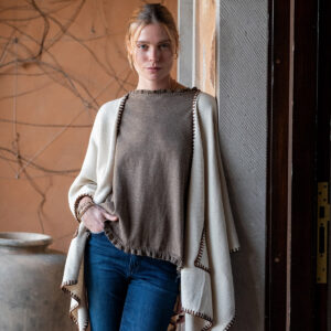 A woman with blonde hair, wearing a taupe top, cream shawl, and blue jeans, stands leaning against a doorway. Large ceramic pots and a tan wall with bare vine branches are visible in the background. Fearrington Village