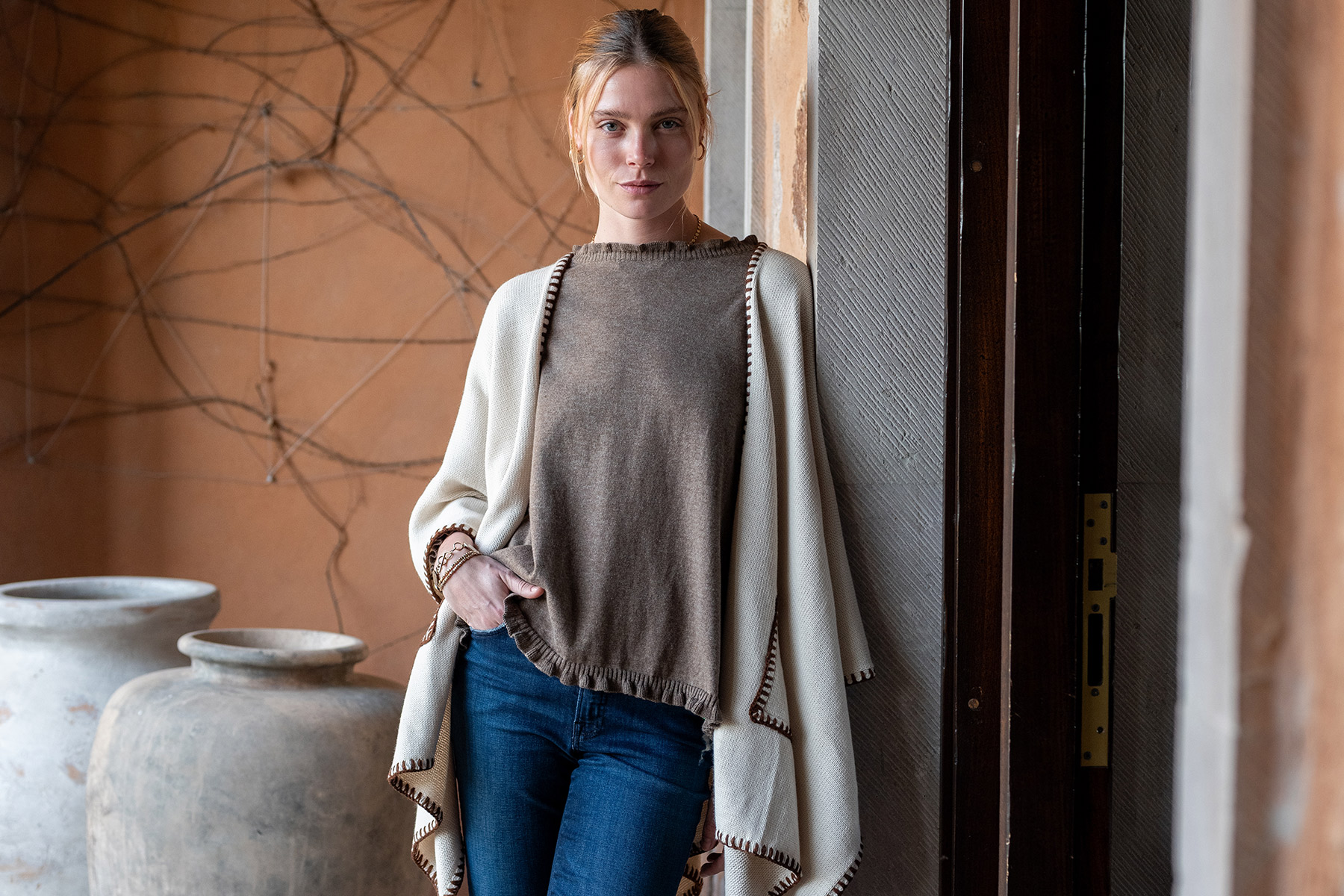 A woman with blonde hair, wearing a taupe top, cream shawl, and blue jeans, stands leaning against a doorway. Large ceramic pots and a tan wall with bare vine branches are visible in the background. Fearrington Village
