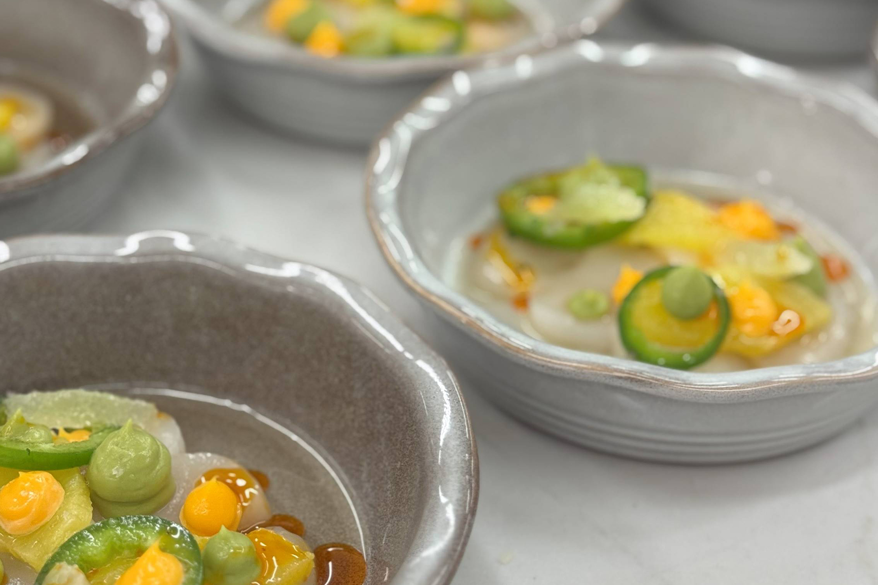 Close-up of several ceramic bowls containing a colorful dish with sliced green vegetables, yellow and orange sauce, and creamy dollops, arranged neatly on a white surface. Fearrington Village
