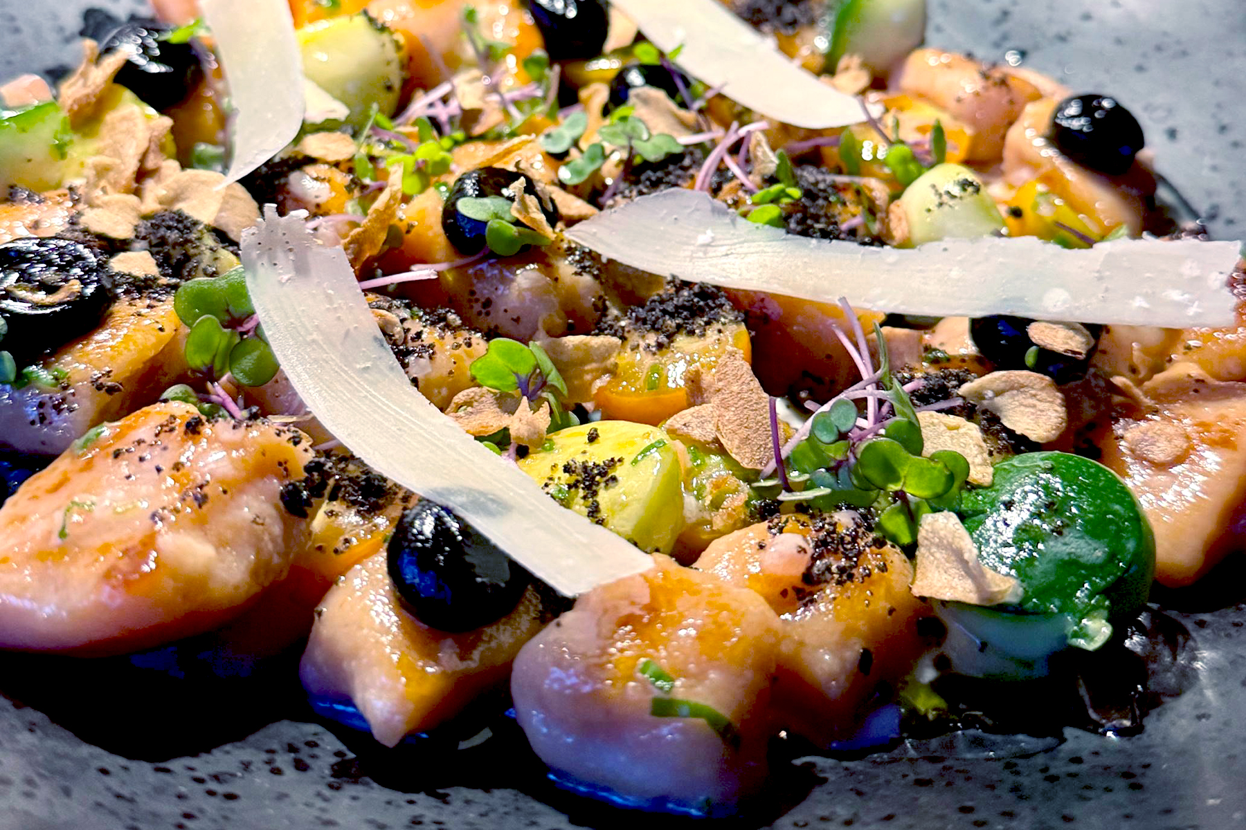 A close-up of a gourmet dish featuring diced salmon, black olive slices, thin white cheese shavings, microgreens, and various colorful garnishes artfully arranged on a dark plate. Fearrington Village