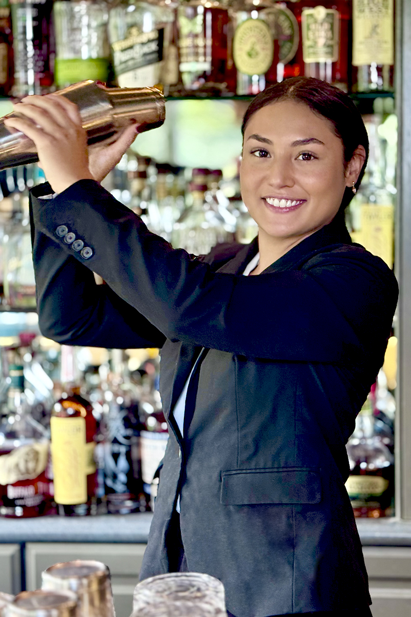 A smiling bartender in a dark blazer shakes a cocktail shaker behind a bar lined with various bottles of liquor and spirits. Fearrington Village