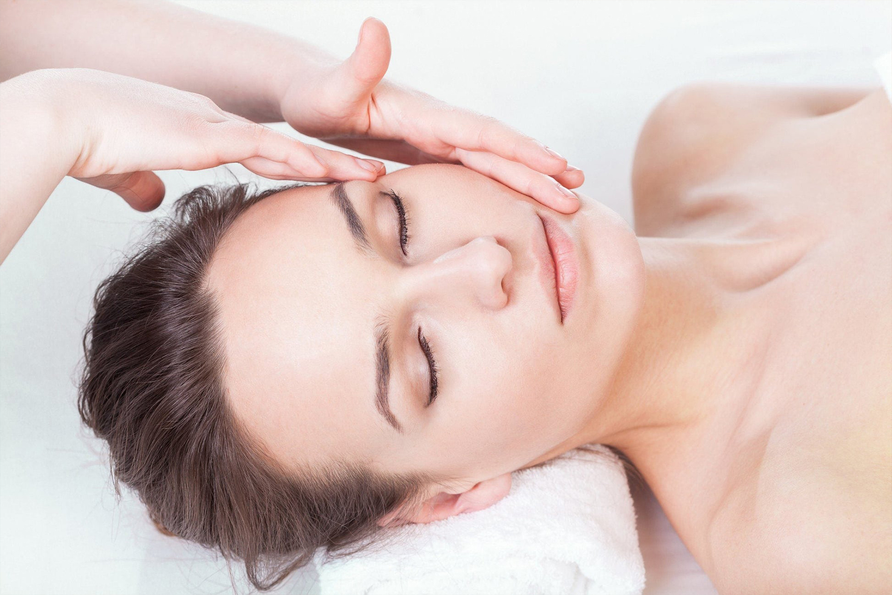 A woman with closed eyes lies on a white towel while receiving a gentle facial massage, with hands softly touching her forehead and cheek. Fearrington Village