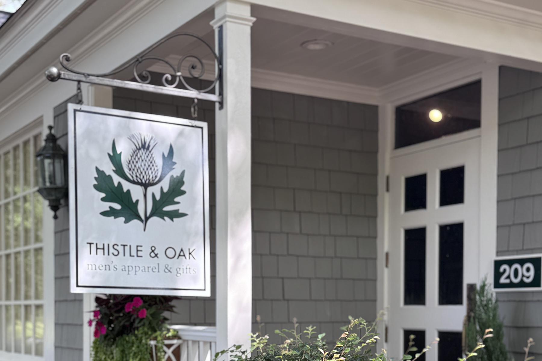 A storefront with gray siding and white trim displays a hanging sign that reads "THISTLE & OAK men's apparel & gifts" with a thistle and oak leaf illustration. The address number 209 is visible next to the door. Fearrington Village