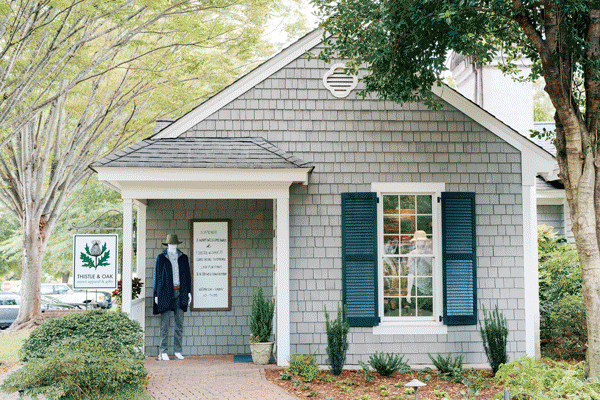 A small gray shingle building with blue shutters and white trim houses a boutique. A mannequin dressed in casual clothes stands near the entrance, and a sign reading “Thistle & Oak” is visible by the walkway. Trees and shrubs surround the shop. Fearrington Village