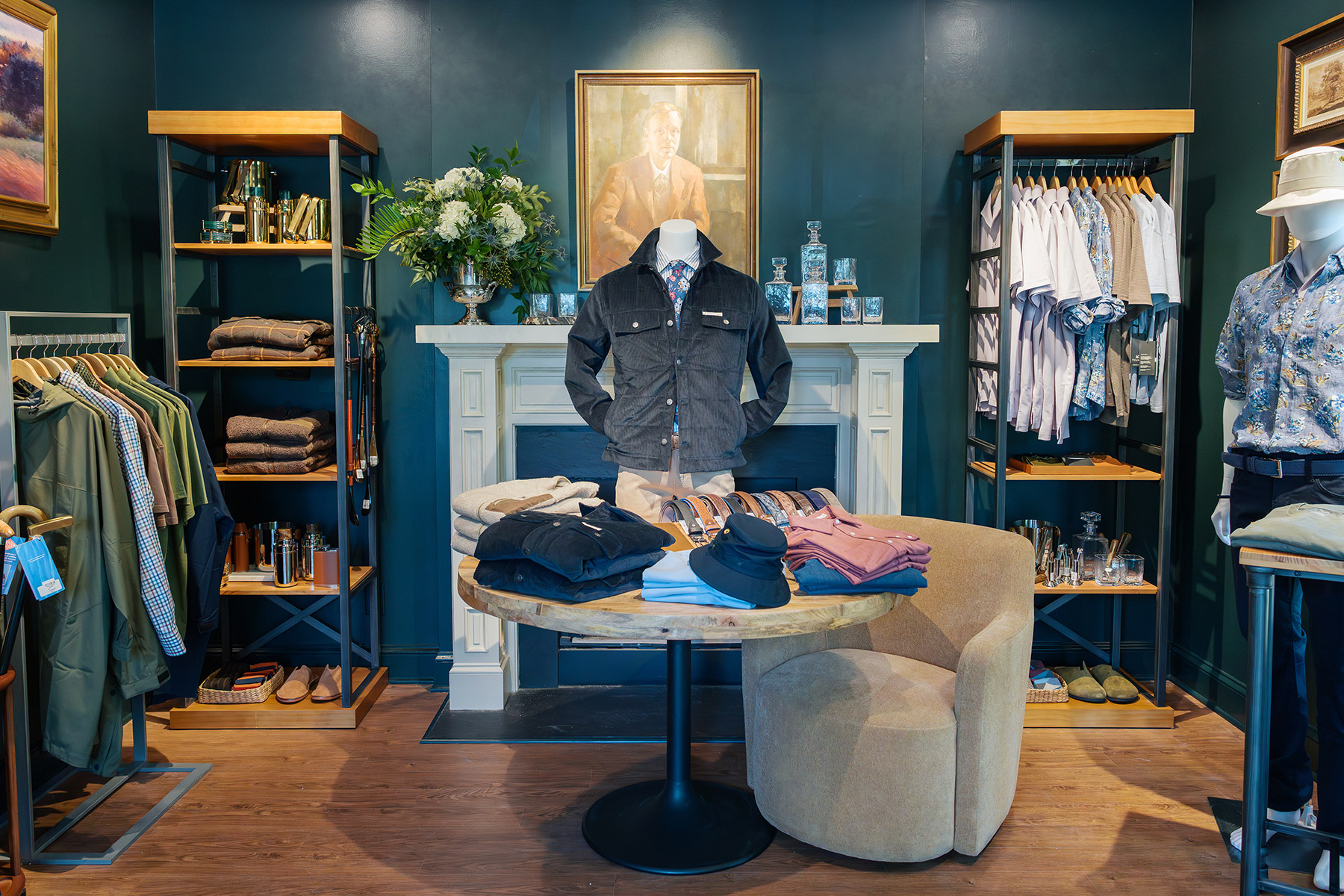 A stylish men's clothing boutique with shirts, jackets, and folded clothes on display. A round table and chair sit in front of a white fireplace with a portrait and flowers above it, surrounded by shelves of garments. Fearrington Village