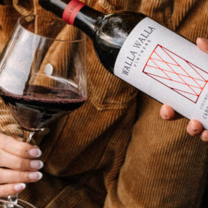 A person holds a glass of red wine while another pours wine from a Walla Walla Vineyards Cabernet Franc bottle. Both wear casual brown clothing. Fearrington Village