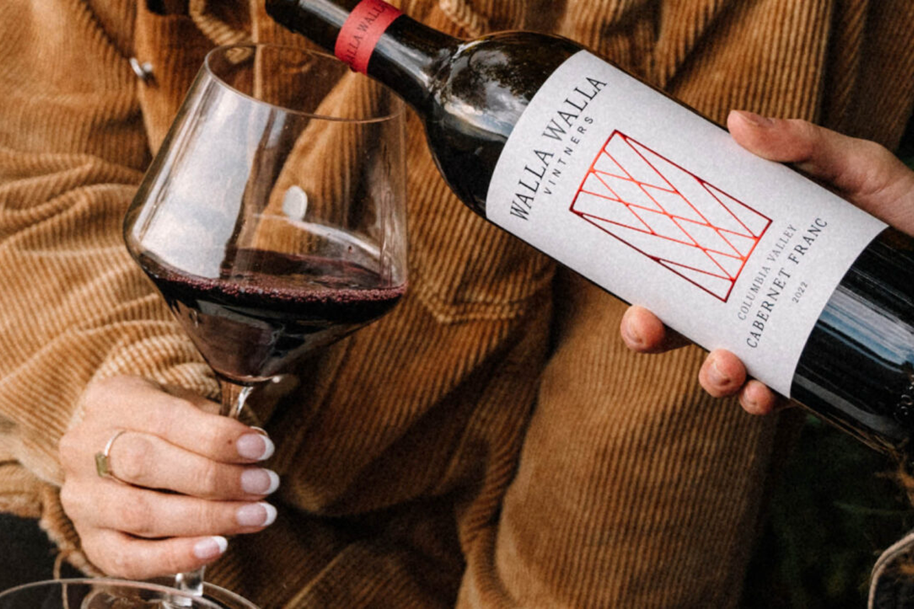 A person holds a glass of red wine while another pours wine from a Walla Walla Vineyards Cabernet Franc bottle. Both wear casual brown clothing. Fearrington Village