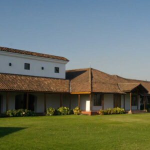 A single-story building with white walls and a brown tiled roof stands beside a well-maintained green lawn under a clear blue sky. Small shrubs line the outside of the building. Fearrington Village
