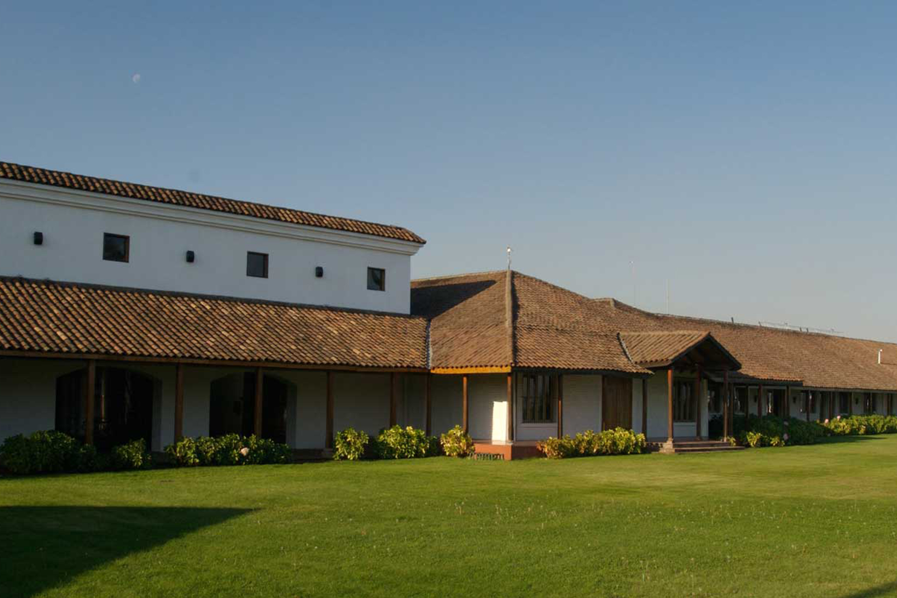 Free Wine Tasting - Casa Lapostolle 31 A single-story building with white walls and a brown tiled roof stands beside a well-maintained green lawn under a clear blue sky. Small shrubs line the outside of the building. Fearrington Village
