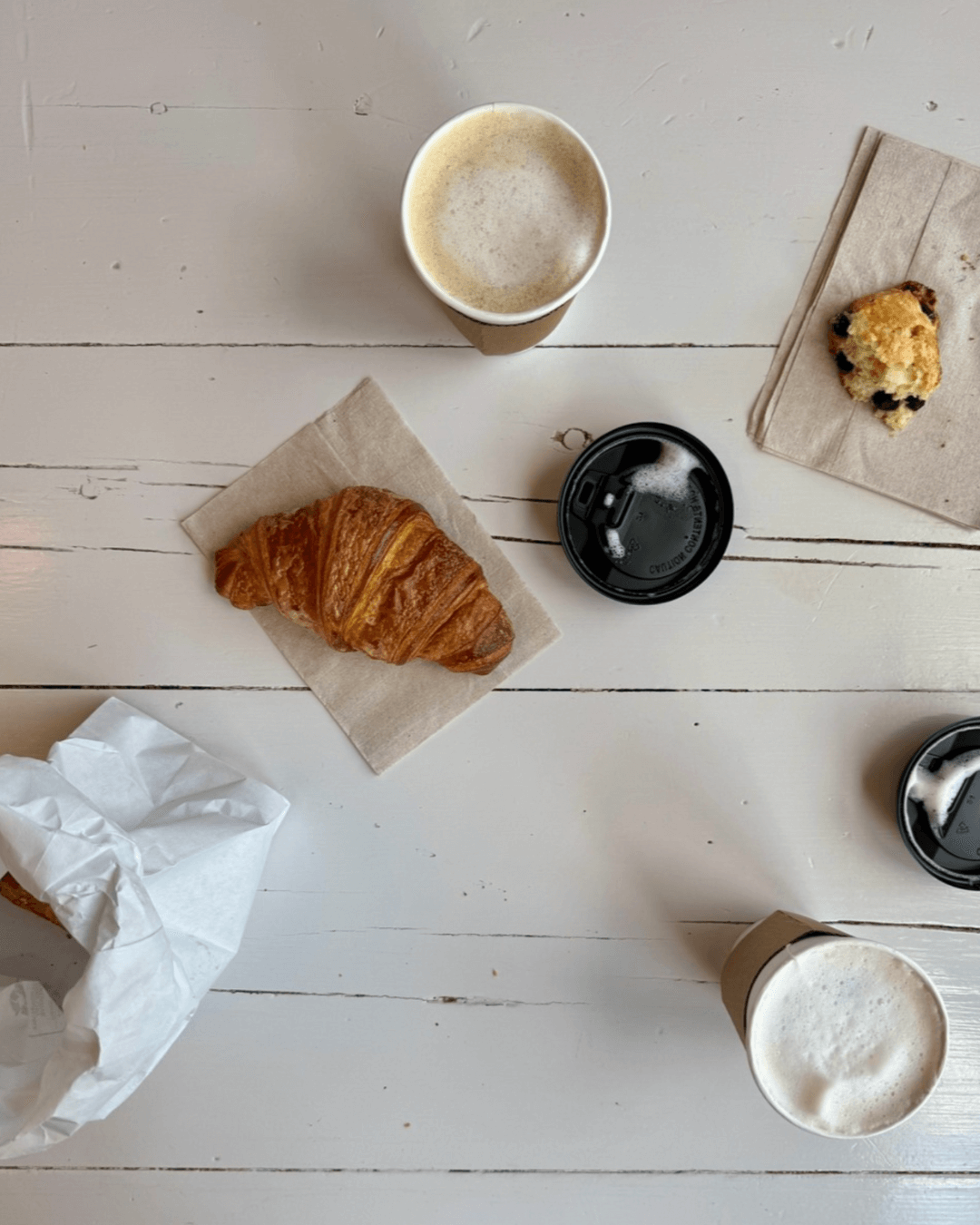 Overhead view of a white table with two cups of coffee, a croissant on a napkin, a scone, a paper bag, and two black coffee cup lids. Fearrington Village