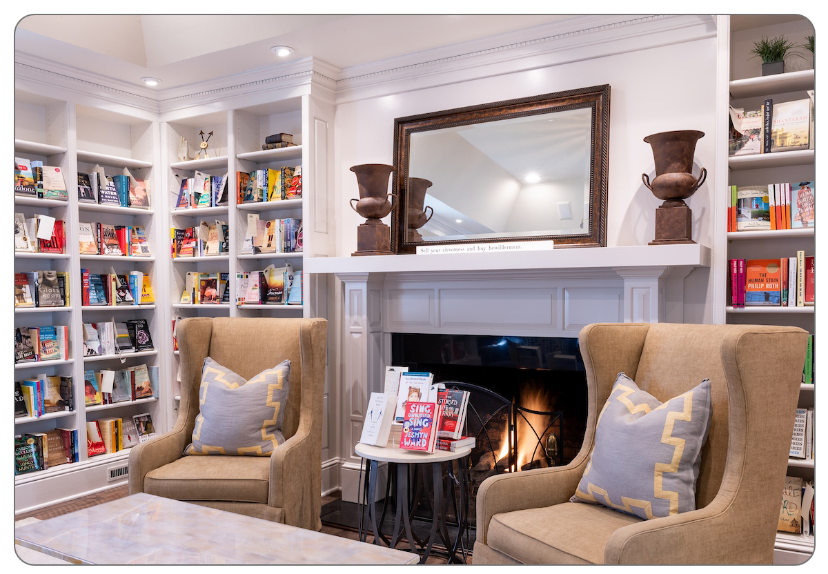 A cozy reading nook with two armchairs, a marble coffee table with books, bookshelves filled with colorful books, and a lit fireplace beneath a large mirror and decorative vases. Fearrington Village