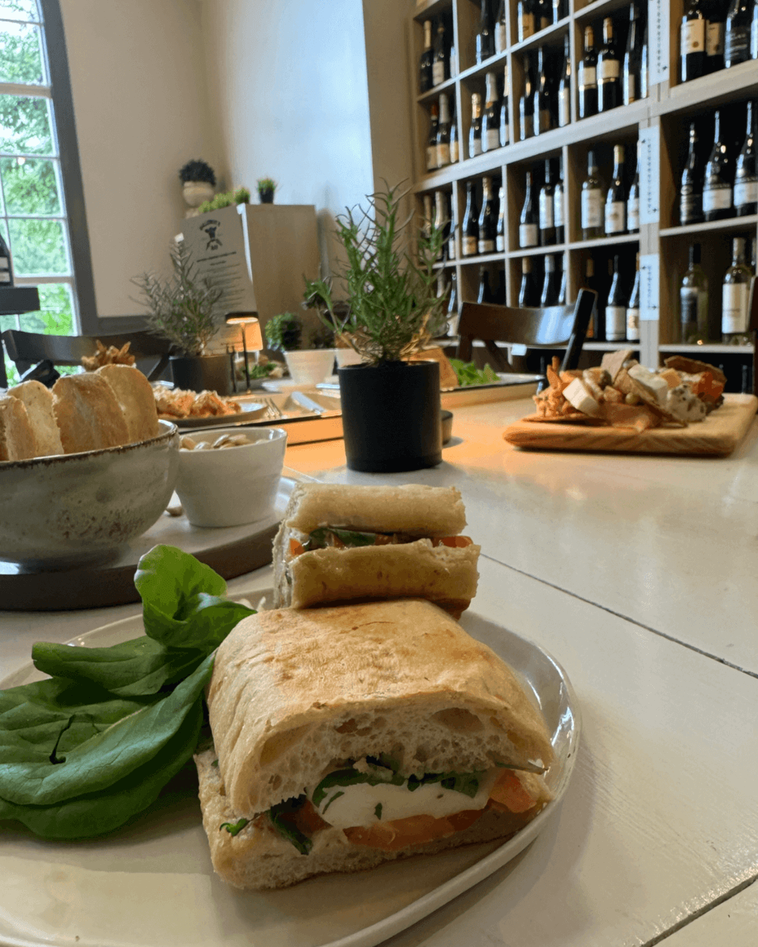 A plate with a ciabatta sandwich filled with mozzarella, tomato, and greens sits on a white table, surrounded by fresh bread, appetizers, and shelves of wine bottles in a cozy, well-lit café. Fearrington Village