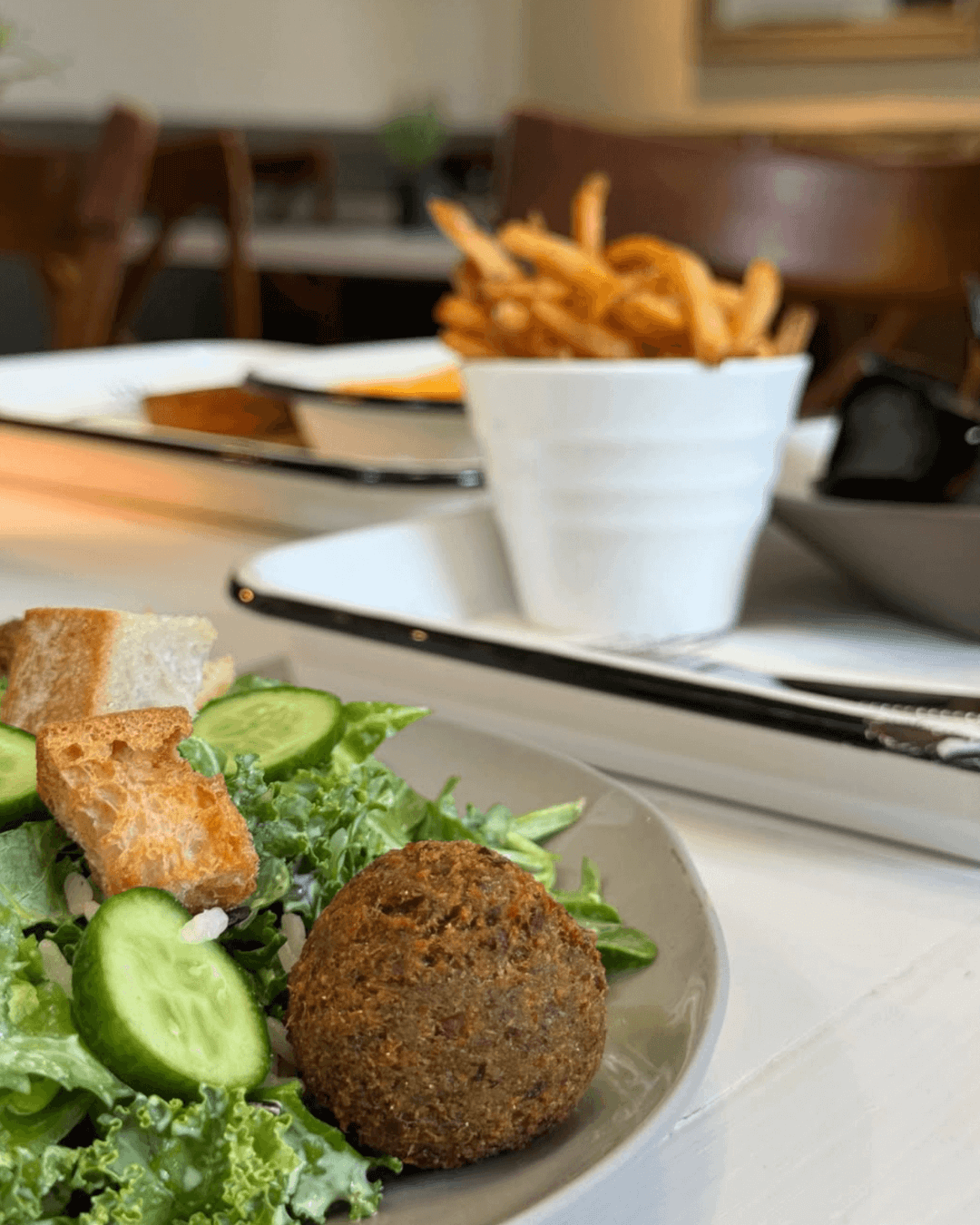 A close-up of a salad with greens, cucumber, croutons, and falafel on a plate, with a bowl of curly fries and another dish blurred in the background on a white table. Fearrington Village