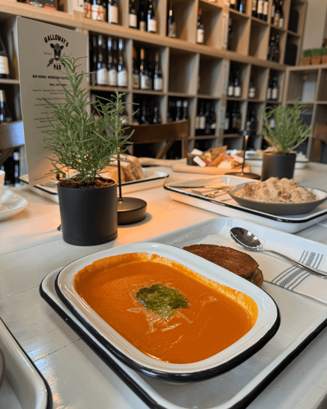 A tray with tomato soup topped with green garnish, a grilled cheese sandwich, and a spoon on a napkin sits on a table in a restaurant with shelves of wine bottles and small potted plants. Fearrington Village