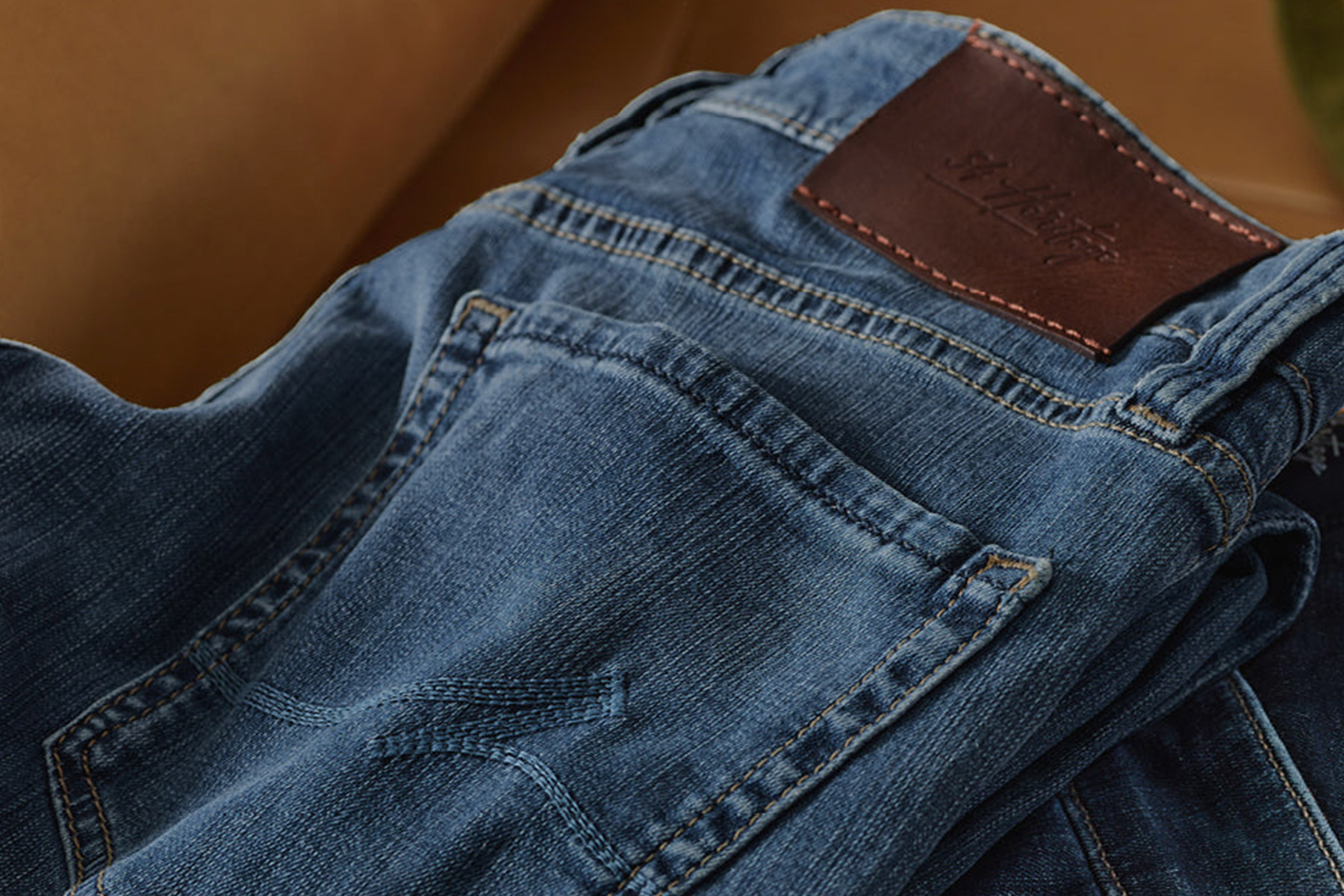 A close-up view of folded blue denim jeans, showing the back pocket and a brown leather patch on the waistband with stitching details. Fearrington Village