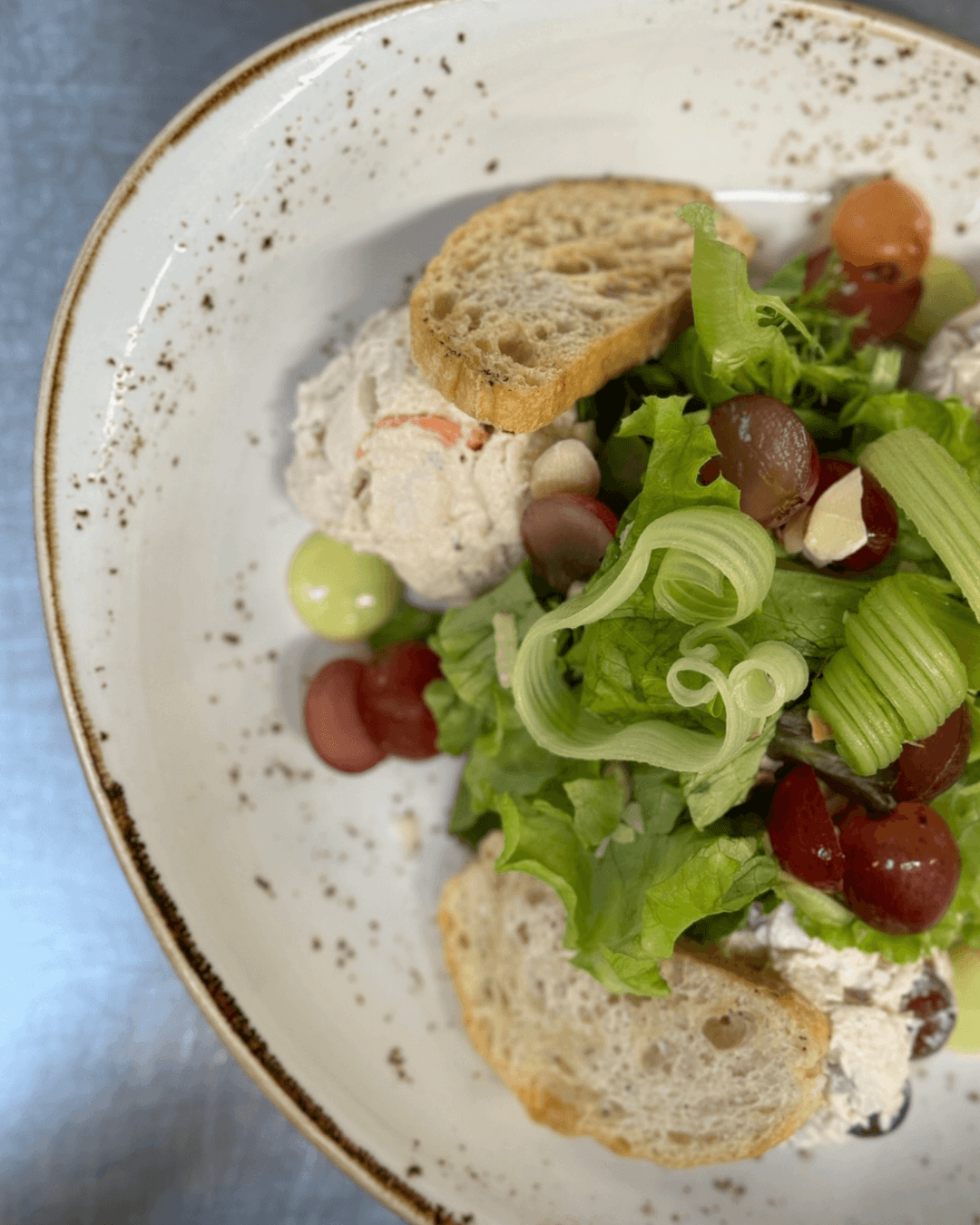 A white plate with chicken salad scoops, leafy greens, sliced grapes, celery ribbons, almond slivers, and two pieces of rustic bread on the side. Fearrington Village