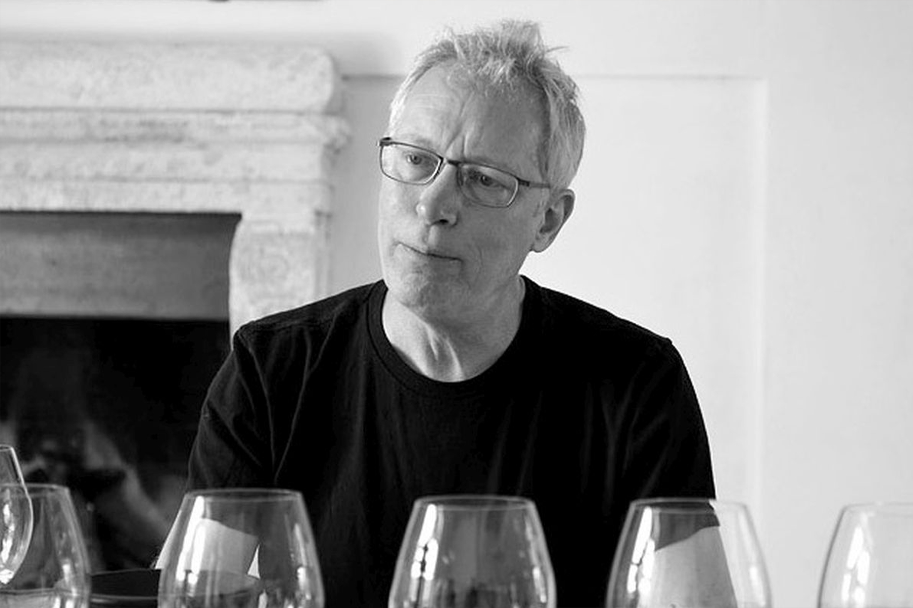 A man with glasses and short, light hair sits at a table with several wine glasses in front of him, looking thoughtful. The image is in black and white. Fearrington Village