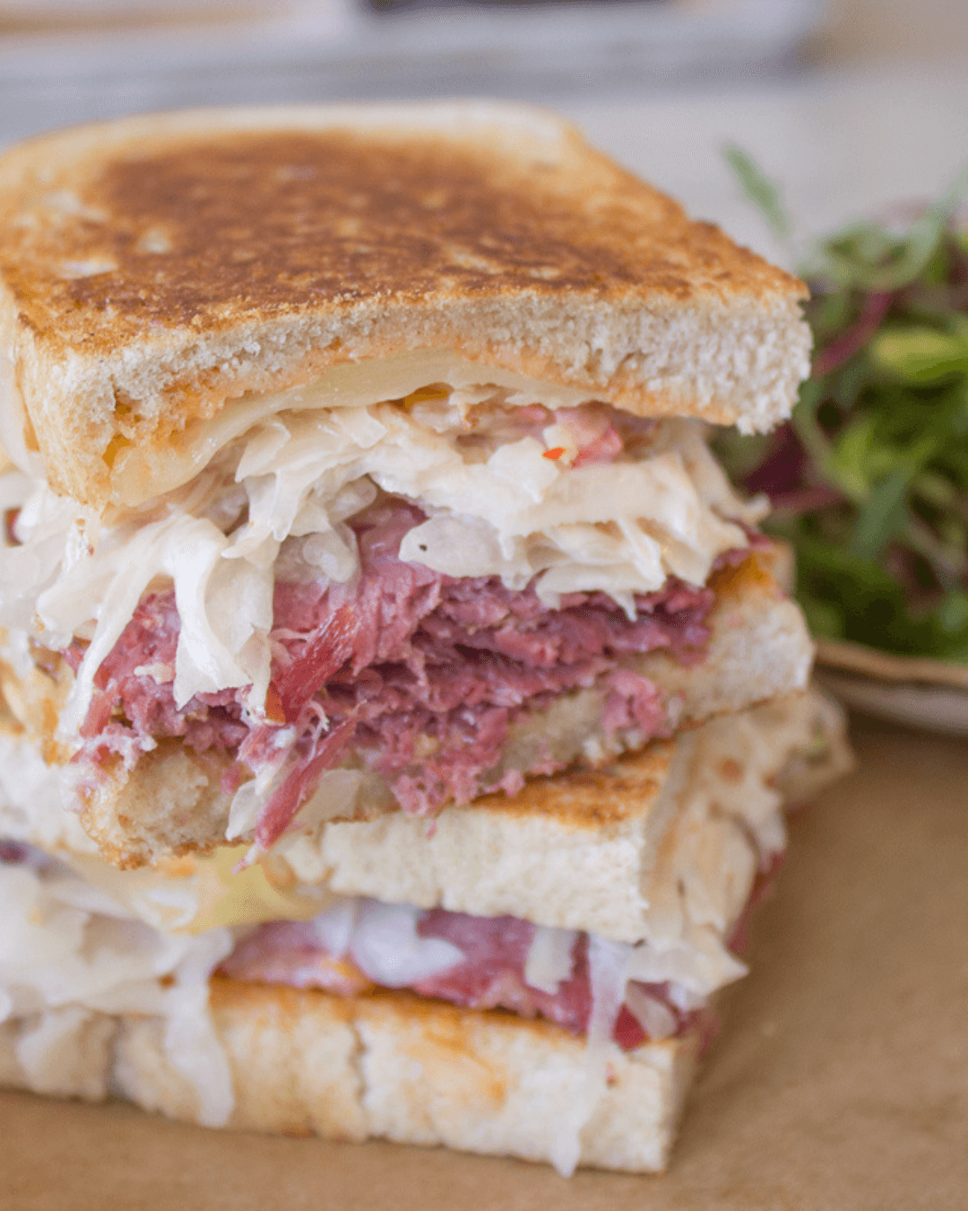 A stacked sandwich with grilled bread, filled with corned beef, sauerkraut, and melted cheese, sits on a brown surface. A small salad with leafy greens is blurred in the background. Fearrington Village