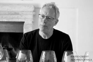 A man with glasses and short light hair, wearing a dark shirt, sits in front of a fireplace with several empty wine glasses in the foreground. Text in the corner reads "Photo Credit: Alfonso Cevola. Fearrington Village