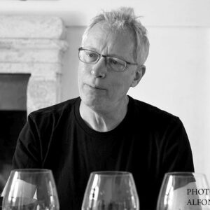 A man with glasses and short light hair, wearing a dark shirt, sits in front of a fireplace with several empty wine glasses in the foreground. Text in the corner reads "Photo Credit: Alfonso Cevola. Fearrington Village