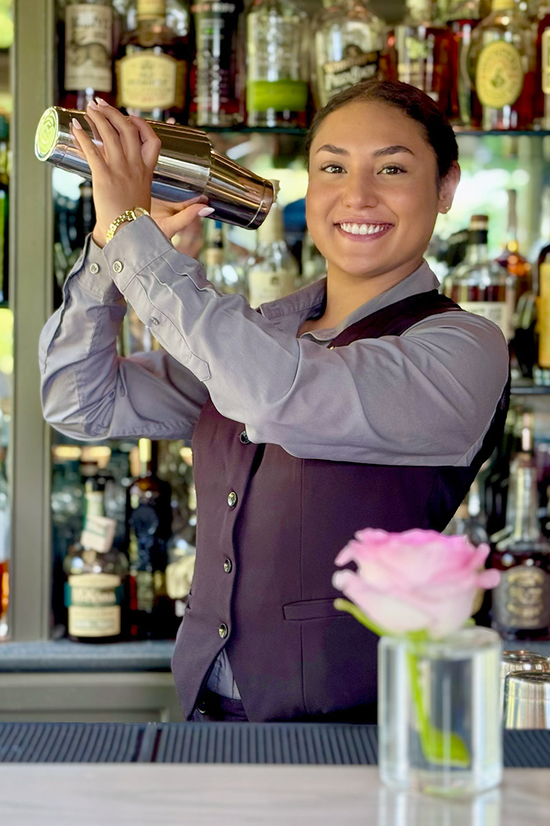 A smiling bartender in a vest shakes a cocktail shaker behind a bar lined with bottles. A pink rose in a glass vase sits on the counter in the foreground. Fearrington Village
