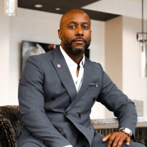 A man in a gray suit sits confidently on a chair, looking at the camera with a calm expression in a modern indoor setting. Fearrington Village
