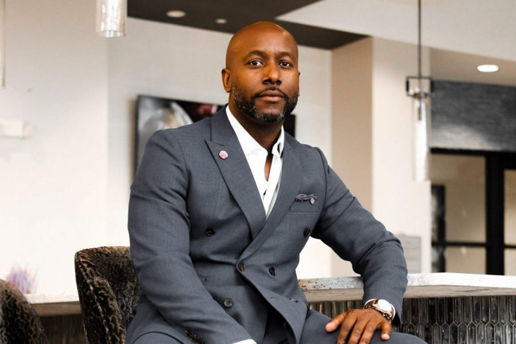 A man in a gray suit sits confidently on a chair, looking at the camera with a calm expression in a modern indoor setting. Fearrington Village