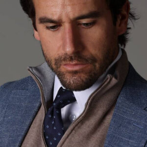 A man with dark hair and a beard wears a blue textured blazer, a beige zip-up sweater, a white shirt, and a navy tie with a subtle pattern. He looks down thoughtfully against a neutral background. Fearrington Village