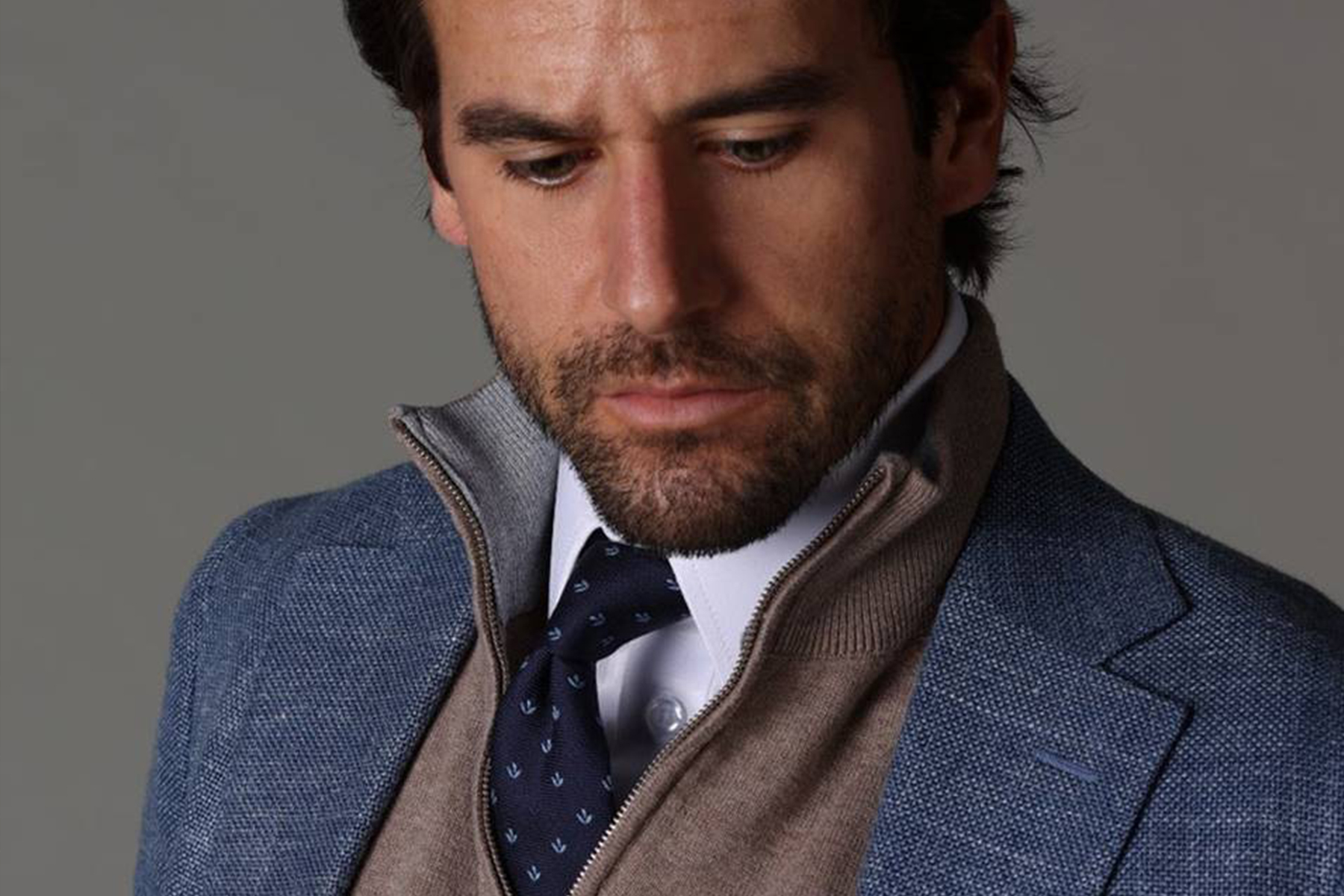 A man with dark hair and a beard wears a blue textured blazer, a beige zip-up sweater, a white shirt, and a navy tie with a subtle pattern. He looks down thoughtfully against a neutral background. Fearrington Village