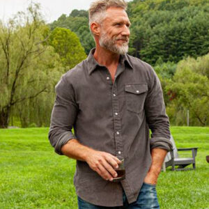 A middle-aged man with gray hair and a beard stands outdoors on green grass, holding a drink and wearing a gray button-down shirt and jeans. Trees, chairs, and a fire pit are visible in the background. Fearrington Village