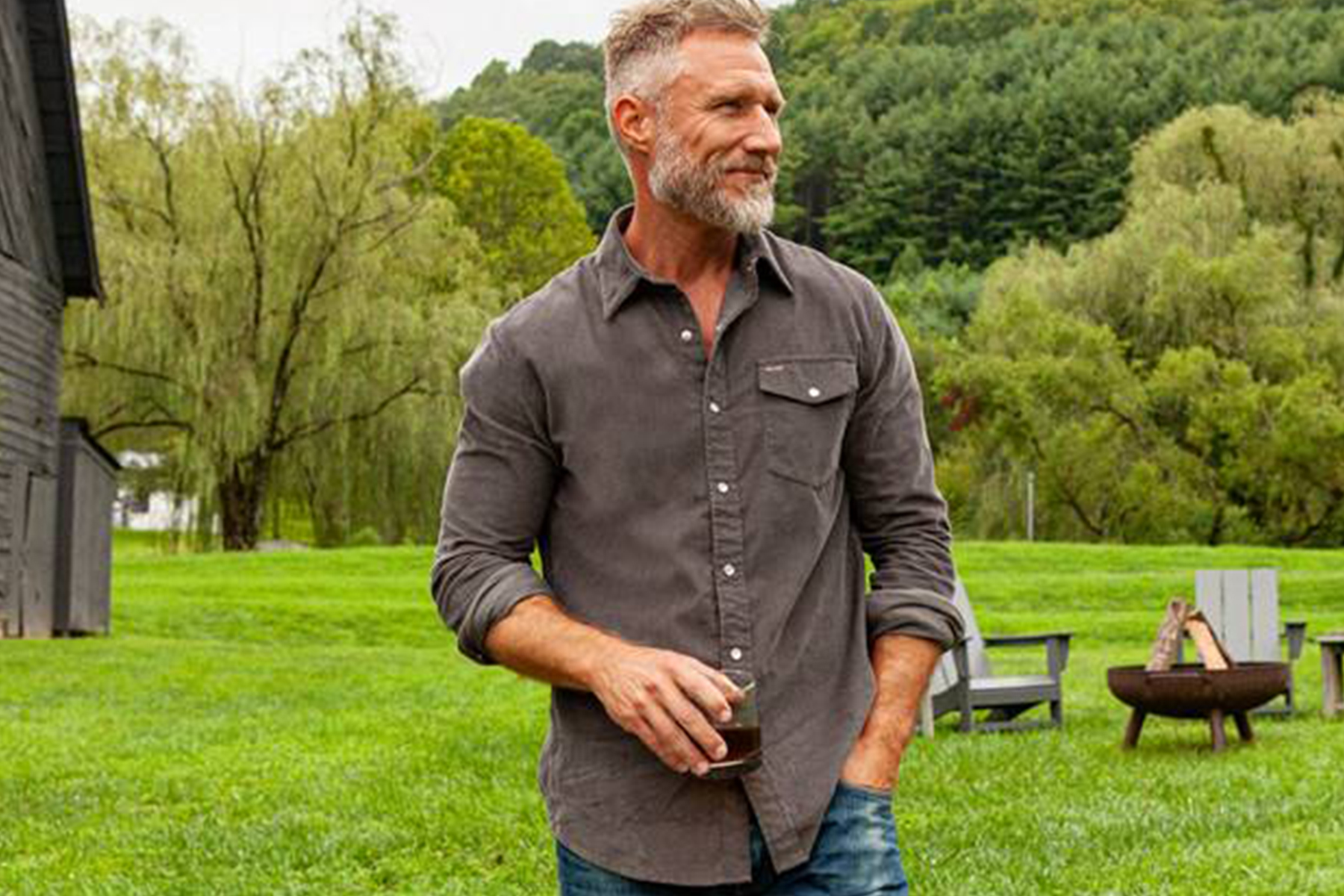 A middle-aged man with gray hair and a beard stands outdoors on green grass, holding a drink and wearing a gray button-down shirt and jeans. Trees, chairs, and a fire pit are visible in the background. Fearrington Village