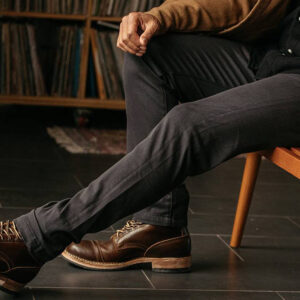 A person wearing dark pants, a brown jacket, and brown leather boots sits on a wooden bench with one leg crossed over the other. A shelf of vinyl records is visible in the background. Fearrington Village