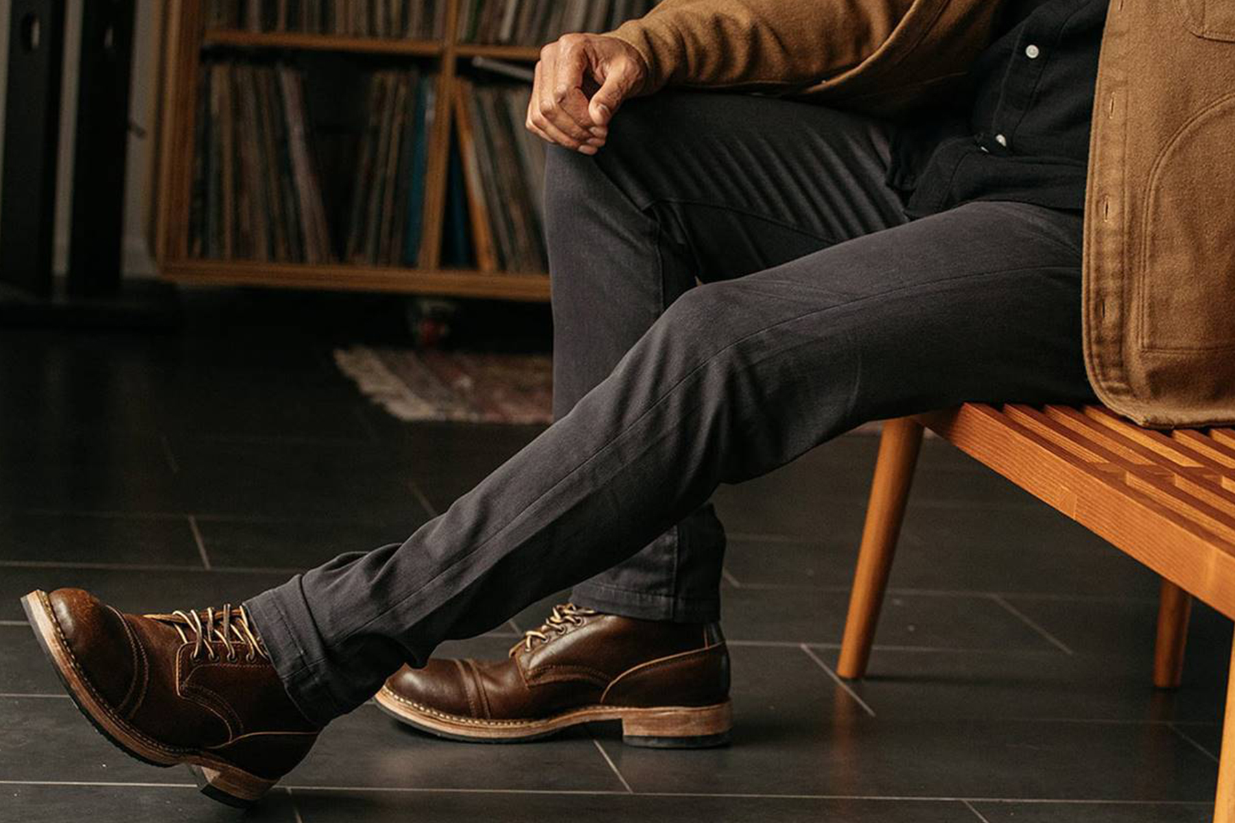 Events 15 A person wearing dark pants, a brown jacket, and brown leather boots sits on a wooden bench with one leg crossed over the other. A shelf of vinyl records is visible in the background. Fearrington Village
