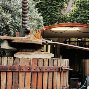 A person operates a large, rustic wooden wine press outside a building covered in green ivy, using a long metal lever to apply pressure. A stainless steel tank is visible inside the open doorway. Fearrington Village