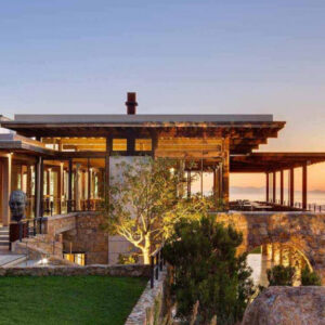 Modern house with stone and glass exterior, large windows, and open terrace overlooking a scenic landscape and ocean at sunset, with green lawn and trees in the foreground. Fearrington Village