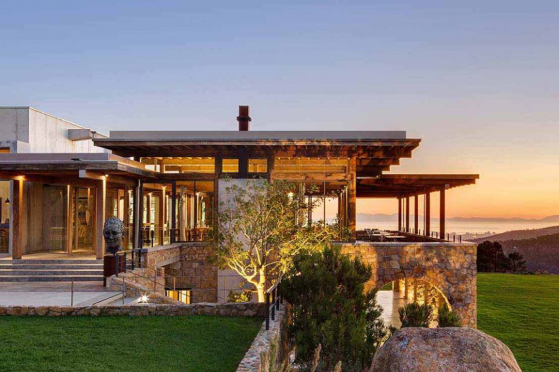 Modern house with stone and glass exterior, large windows, and open terrace overlooking a scenic landscape and ocean at sunset, with green lawn and trees in the foreground. Fearrington Village