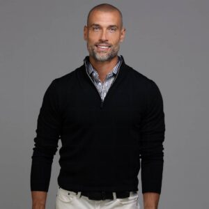 A man with a shaved head and beard, wearing a black sweater over a checkered shirt and white pants, stands smiling against a plain gray background. Fearrington Village