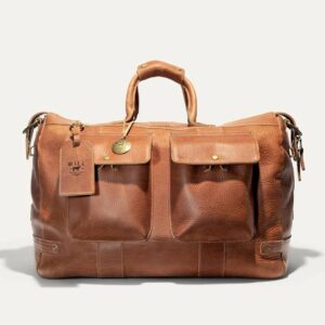 A large brown leather duffel bag with two front pockets, a top handle, and a luggage tag attached. The bag has brass hardware and a textured finish. Fearrington Village