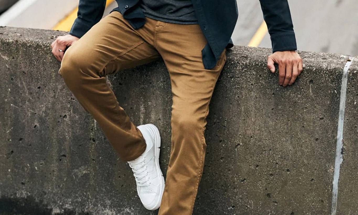 Thistle & Oak Fall 2025 Trunk Show: DU/ER 1 A person wearing tan pants, a dark jacket, and white sneakers sits casually on a concrete barrier with one leg bent and one hand resting on the barrier. Fearrington Village