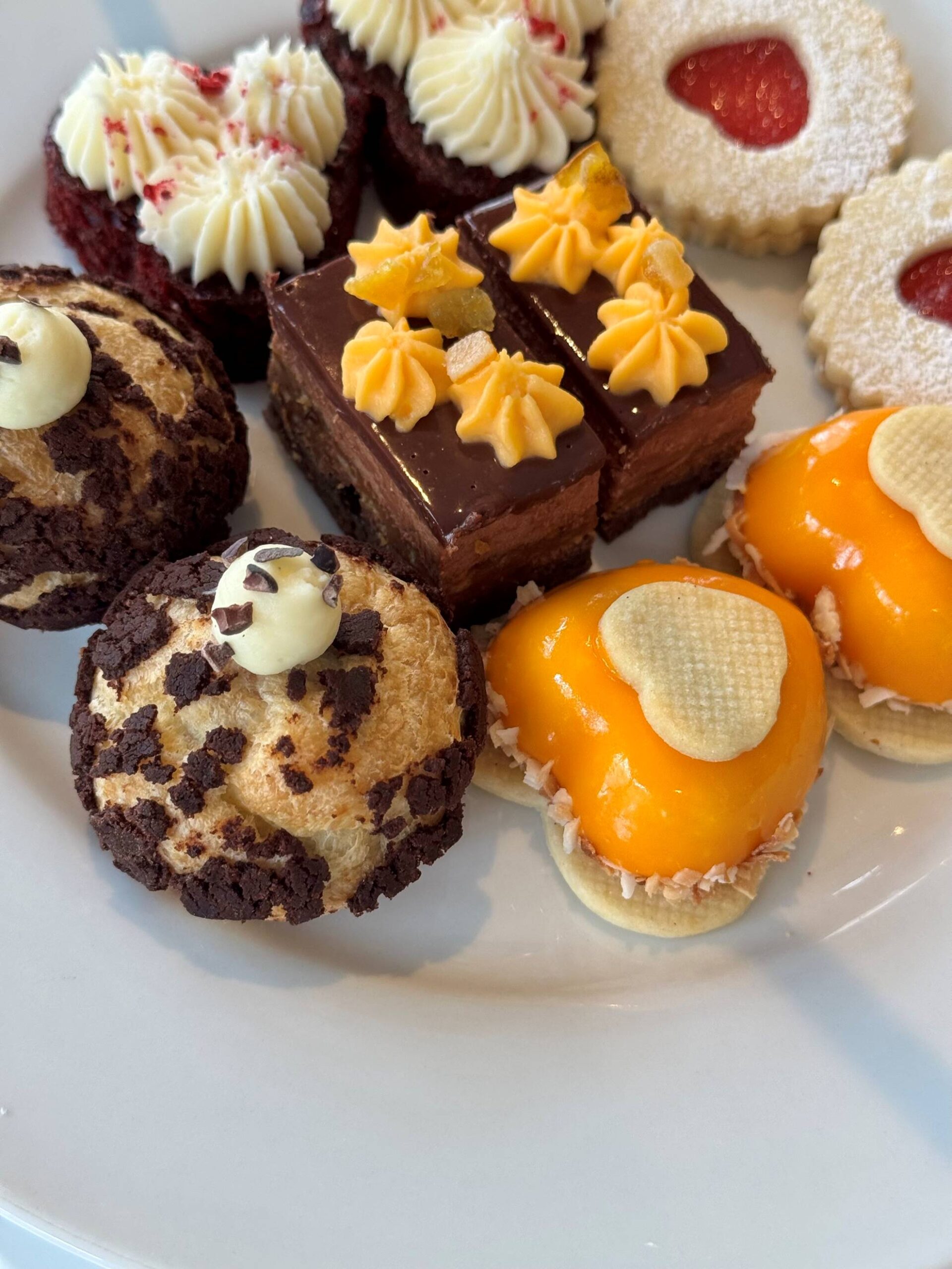 A plate of assorted pastries, including chocolate cream puffs, chocolate layered cakes with yellow topping, red velvet mini cakes with cream, heart-shaped cookies with jam, and orange-glazed tarts. Fearrington Village