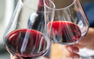 Two people clinking glasses of red wine together in a celebratory toast, with a blurred background and natural light coming from a window. Fearrington Village
