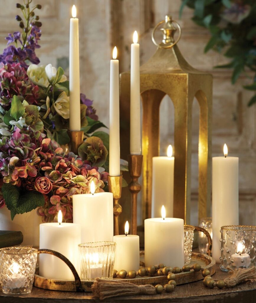 A display of lit white candles of various heights, surrounded by glass candle holders, gold beads, a gold lantern, and a bouquet of purple and green flowers on a rustic wooden surface. Fearrington Village